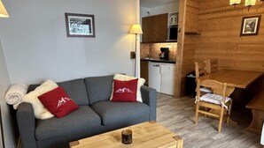 A cozy living area with a grey sofa, two red pillows, a wooden coffee table, and a wooden dining table with chairs. A kitchen with wooden cabinets and a microwave is visible in the background. - 130m from ski slopes, 8th floor, view ski slopes, balcony, ski locker, 33m², Méribel-Mottaret (Méribel - Les Allues)