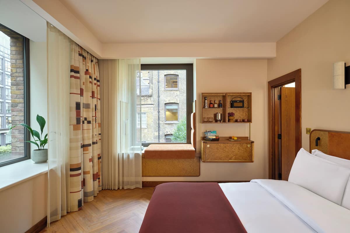 Habitación de hotel con una cama grande, una ventana con cortinas, una zona de descanso pequeña y un estante con artículos de aseo.