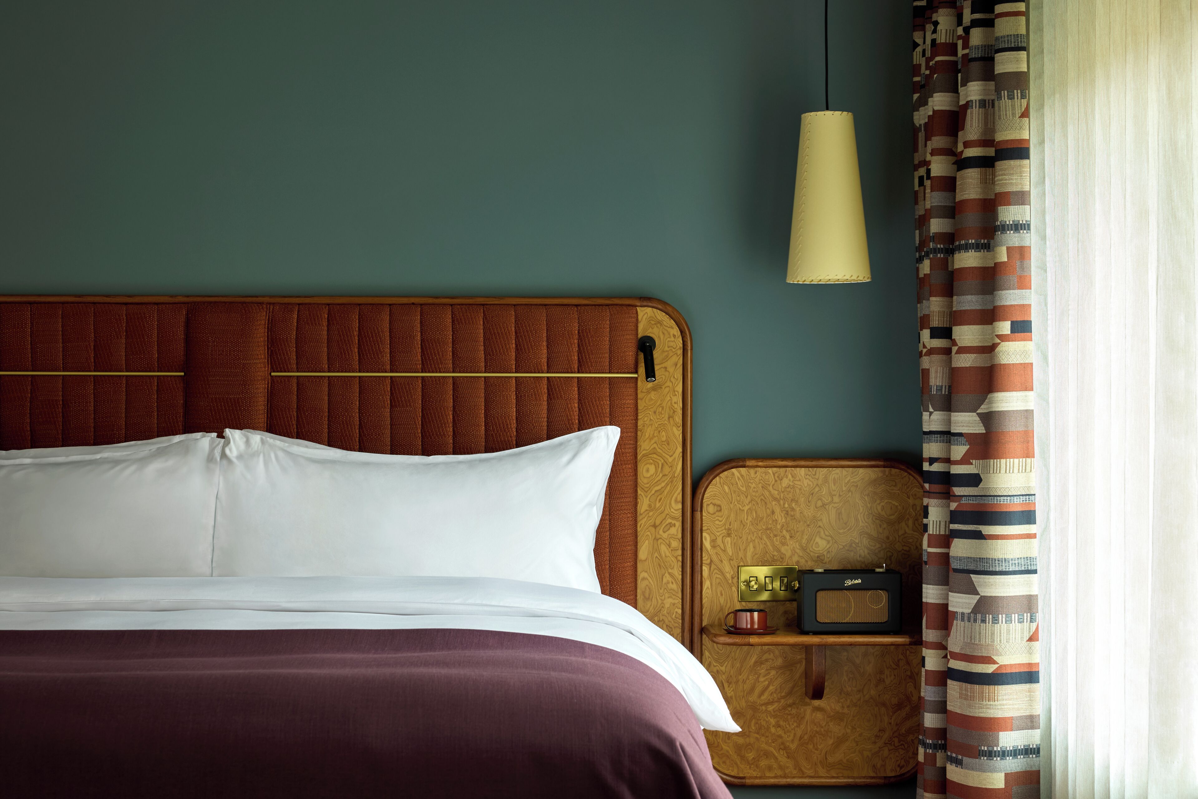 A neatly made bed with a wooden headboard, a bedside table with a radio, and a patterned curtain.