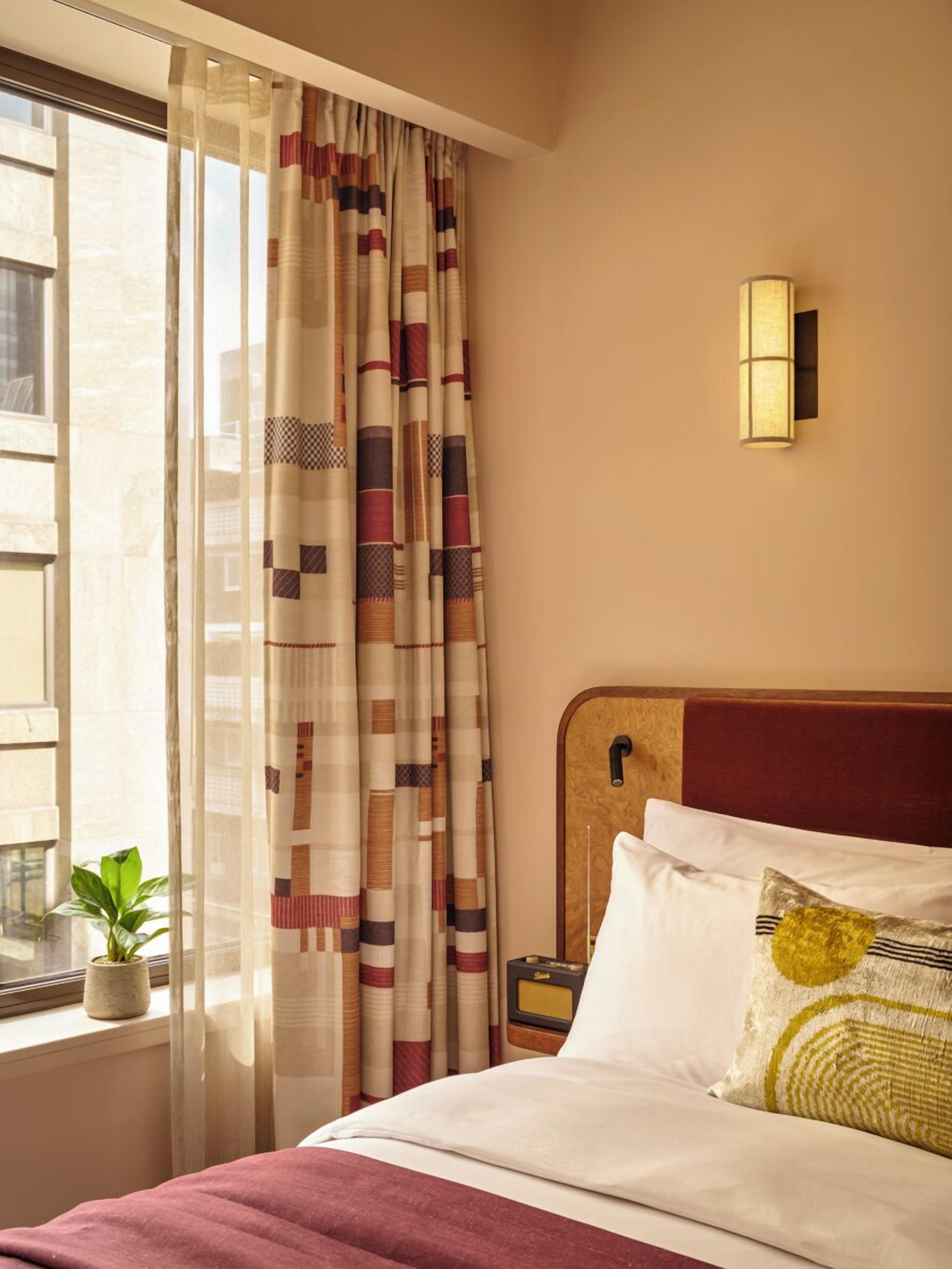 Ein Hotelzimmer mit Bett, einem fenster mit Vorhängen, einer Wandleuchte und einer kleinen Pflanze auf der Fensterbank.