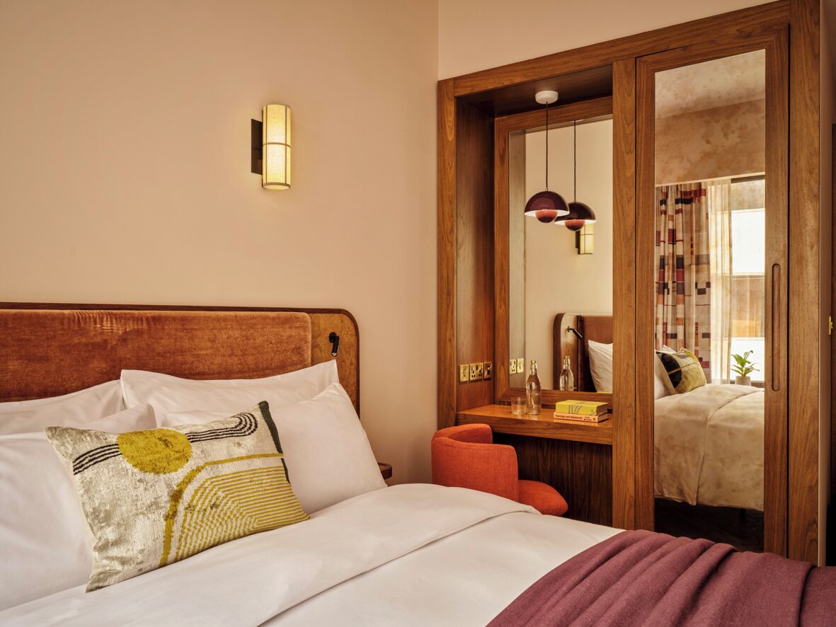 Una habitación de hotel con cabecera de madera, cama con ropa de cama blanca, espejo y mesita de noche de madera.