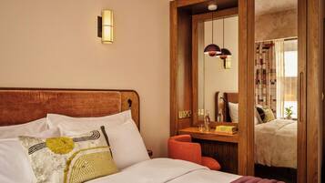 Una habitación de hotel con cabecera de madera, cama con ropa de cama blanca, espejo y mesita de noche de madera.