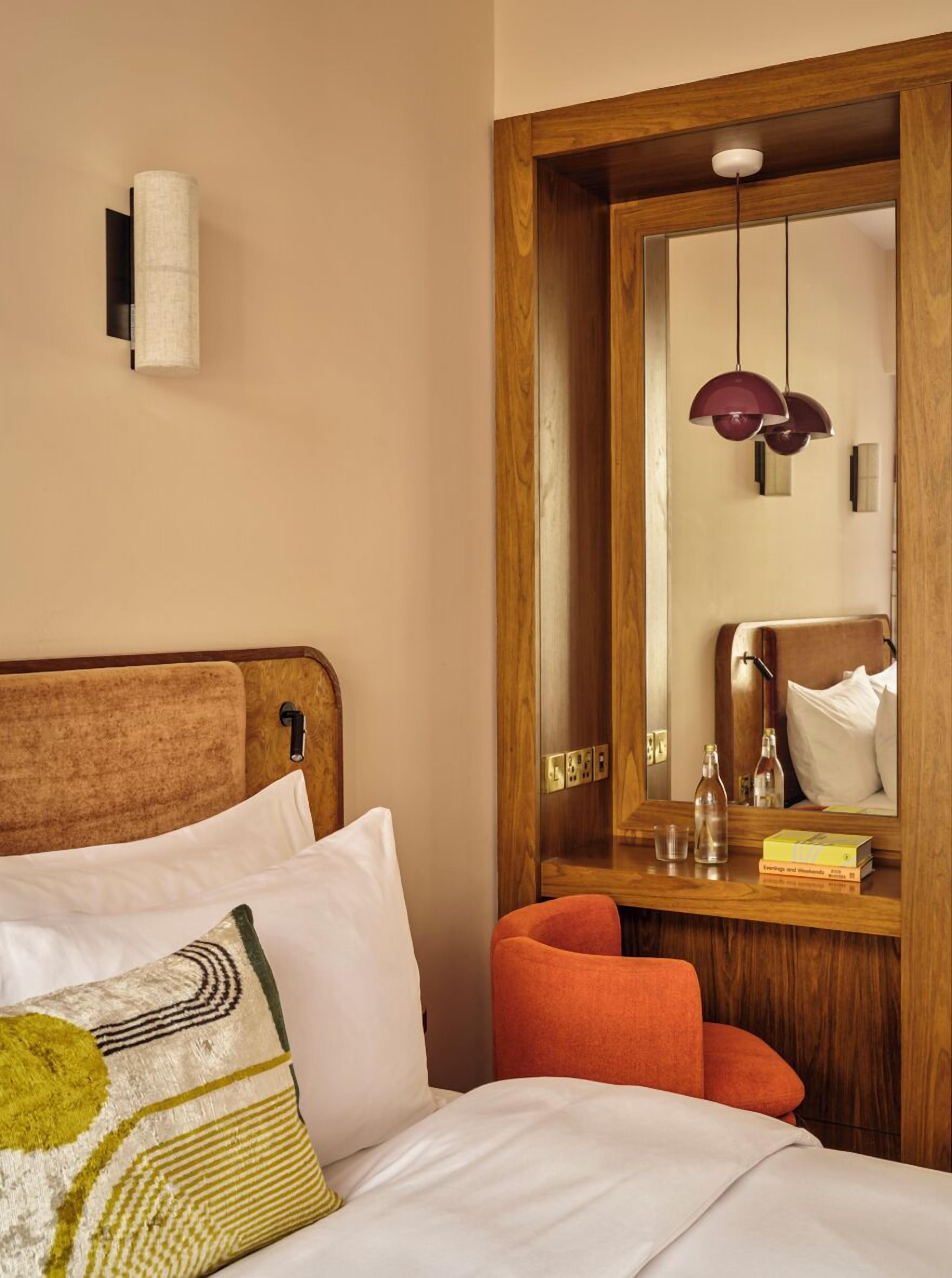 A hotel room with a wooden headboard, a bed with white and patterned pillows, a wooden nightstand with a mirror, a red chair, and a small table with books and bottles.