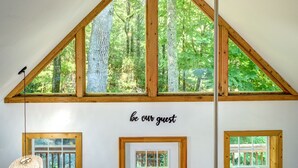 A room with a sloped ceiling, wooden beams, and large windows offering a view of a forest. A sign on the wall reads "be our guest." - Visit #mytreetophideaway located in Banner Elk, NC, nestled high in the trees.  (Banner Elk)