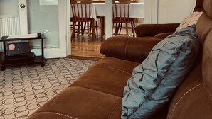 A living room with a brown sofa, a dining table with chairs, and a kitchen area visible through an open door.
