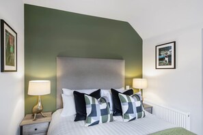 A neatly made bed with a grey headboard, two bedside tables with lamps, and two framed pictures on the walls. - The Woodside Apartments (Doune)