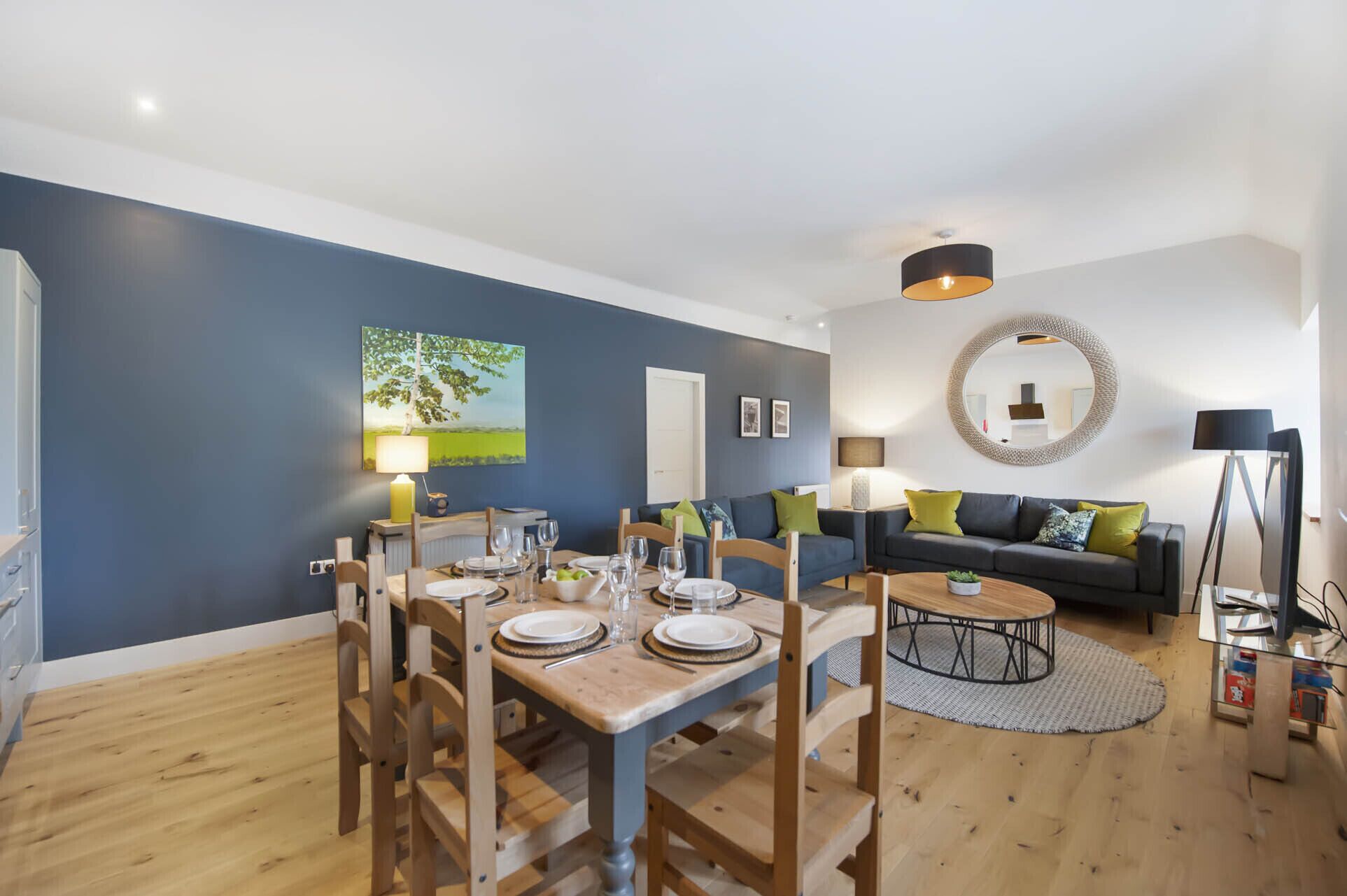 Sala de estar moderna com área de jantar, apresentando uma parede azul, piso de madeira e uma mesa de centro redonda.