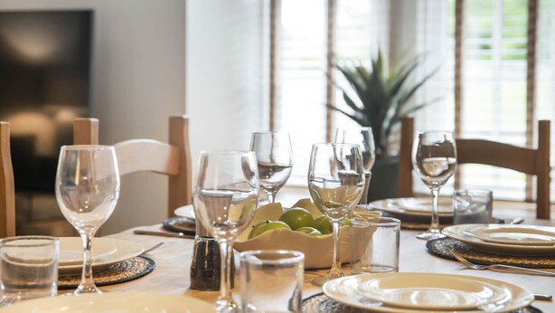 A dining table set with plates, glasses, and a bowl of fruit, in a room with large windows and a plant in the background. - The Woodside Apartments (Doune)