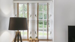 A room with a window featuring horizontal blinds, a floor lamp on a tripod stand, and two small potted plants on a shelf below the window. - The Woodside Apartments (Doune)