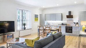 A modern living room with a grey sofa, a dining table with chairs, a TV, and a large window with blinds. - The Woodside Apartments (Doune)