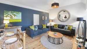 A modern living room with two sofas, a coffee table, and a dining area with a table set for four. - The Woodside Apartments (Doune)