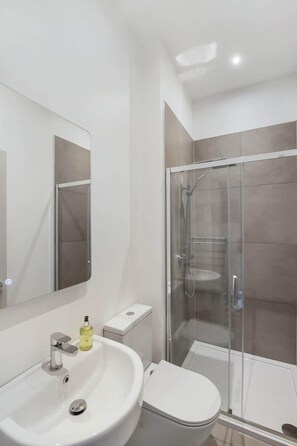 A modern bathroom with a white sink, mirror, toilet, and glass-enclosed shower. - The Woodside Apartments (Doune)