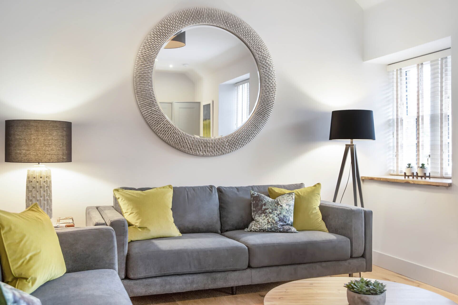 A living room with a grey sofa, yellow and grey cushions, a round mirror, a floor lamp, and a small potted plant.