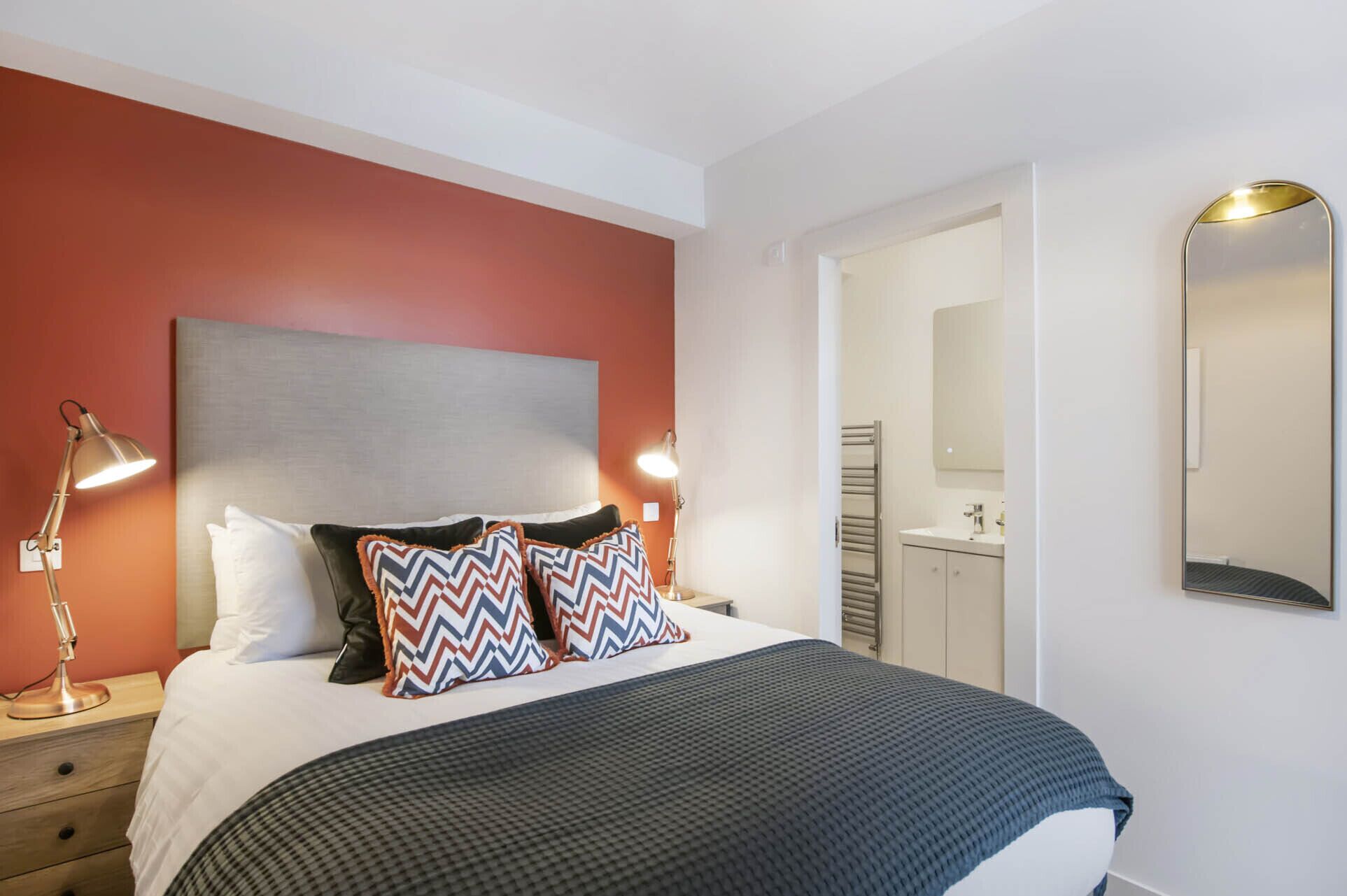 Una habitación de hotel con una cama, lámparas de noche, un espejo y un baño visible a través de una puerta abierta.