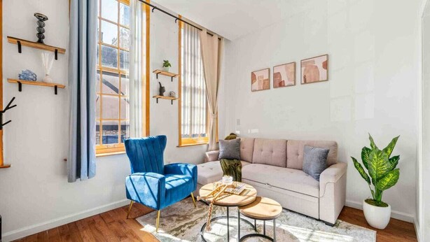 Ein modernes Wohnzimmer mit blauem Sessel, beigefarbenem Sofa und Couchtisch. Regale mit Dekorationselementen und ein großes Fenster mit Vorhängen.
