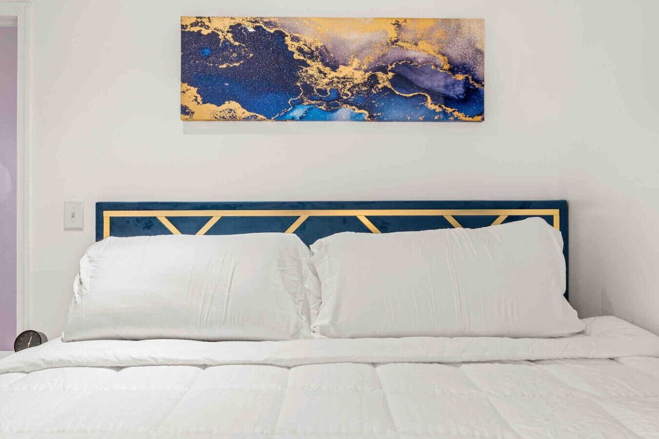 A bed with a blue and gold headboard, white bedding, and a large abstract painting above it.