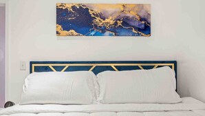 Ein Bett mit blau-goldenem Kopfteil, weißer Bettwäsche und einem großen abstrakten Gemälde darüber.