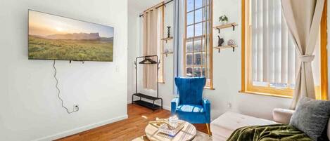 A living room with a large window, a blue chair, a sofa, a coffee table, and a TV mounted on the wall.