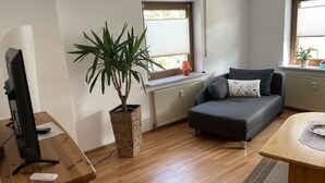 Una sala de estar moderna con piso de madera, un sofá gris, una mesa de madera, un televisor, una planta en maceta y una ventana con persianas.