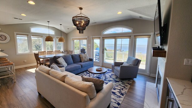 A spacious living room with a large sofa, a coffee table, and a television. There are large windows providing a view of the outdoors. - Amazing Sunsets & Luxury - Soundfront Pet Friendly Oasis w/ Elevator & Hot Tub (Kitty Hawk)