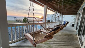 A wooden porch with a swing chair and a view of the sunset over the water. - Amazing Sunsets & Luxury - Soundfront Pet Friendly Oasis w/ Elevator & Hot Tub (Kitty Hawk)