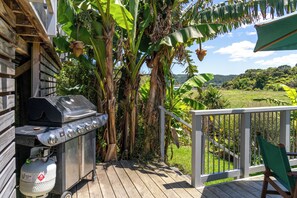 Une terrasse en bois équipée d’un barbecue et d’une bouteille de gaz, entourée de bananiers et offrant une vue sur un paysage verdoyant.