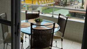 A balcony with a glass table and wicker chairs, overlooking a street with palm trees and buildings. - Apto Maravilhoso com Vista Para o Mar, 50mts da Orla da Praia Grande em Ubatuba (Praia Grande Ubatuba)