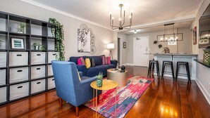 A modern living room with a blue sofa, a coffee table, and a colorful rug. There is a bookshelf with storage bins and a plant. A kitchen area with a bar stool and a TV is visible in the background. - Downtown 2BR King Bed FREE Parking 1413 (Atlanta)