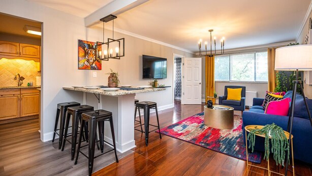 A modern kitchen with a central island, bar stools, a dining area with a colorful rug, a living room with a blue sofa and yellow pillows, and a large window with curtains. - Downtown 2BR King Bed FREE Parking 1413 (Atlanta)