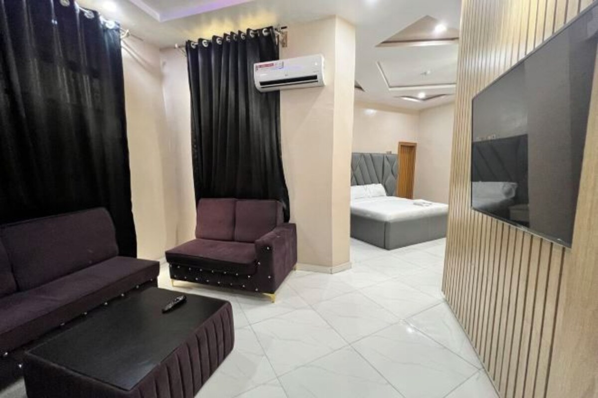 A modern hotel room with a sofa, a small armchair, a flat-screen TV mounted on the wall, and an air conditioning unit.