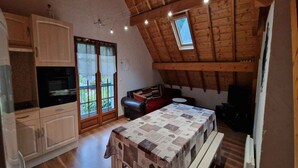 A cozy attic room with a wooden ceiling, a dining table with a quilt, a sofa, and a kitchen area with cabinets and a stove.