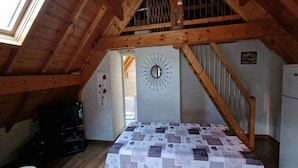 A bedroom with a wooden ceiling, a bed with a patterned bedspread, a wall-mounted mirror, and a small painting.