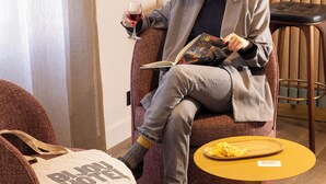 Une personne est assise sur une chaise marron, en train de lire un livre, et tient un verre de vin rouge à la main. Un sac en toile beige est posé sur le sol, à côté de la chaise.