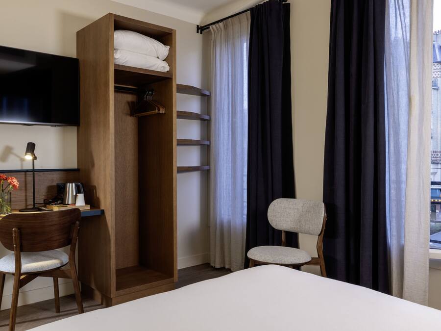 Una camera d'albergo con un letto, una scrivania con una sedia, un armadio, una TV e una finestra con tende.