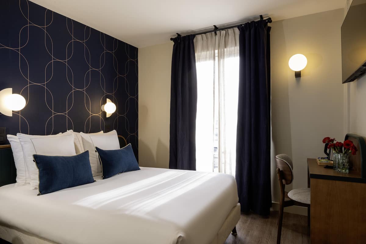 A hotel room with a large bed, a desk with a chair, a vase with flowers, and a window with curtains.