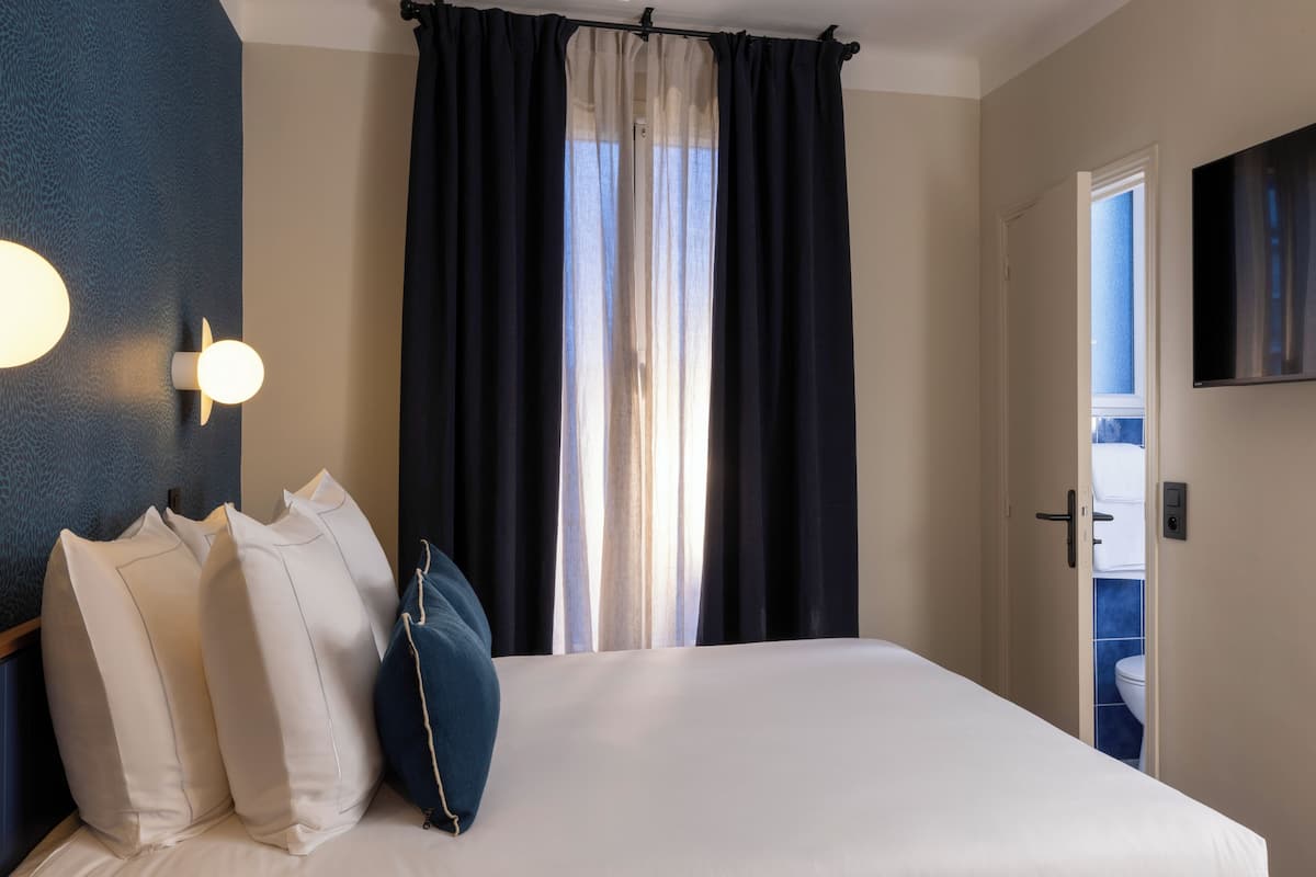 A hotel room with a bed, pillows, a TV, and a window with curtains.