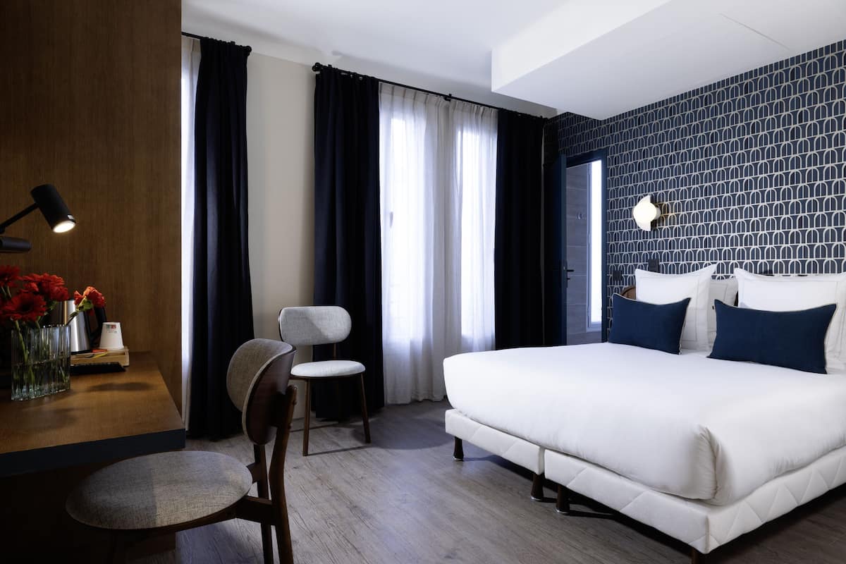 A modern hotel room with a large bed, a desk with a lamp, a chair, and a view of the city through the window.