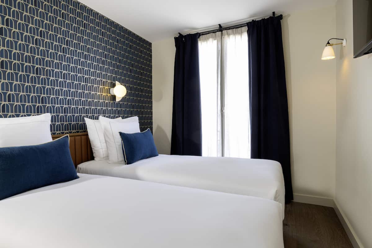 A hotel room with two beds, a patterned wall, a window with curtains, and a wall-mounted light.