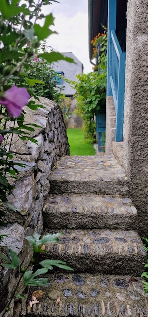 Un escalier en pierre mène à un bâtiment doté de balustrades bleues, entouré de verdure et d’un mur de pierre.
