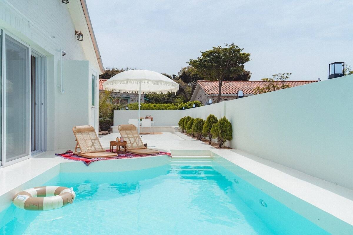 A private pool with lounge chairs and a sun umbrella, surrounded by a white wall and a glass door.