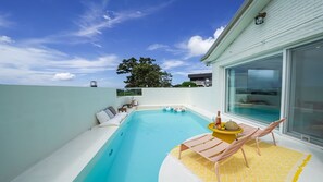 A modern outdoor pool area with a glass-enclosed room, a lounge chair, and a table with a drink and fruit. - Seonheul an (Jeju City)