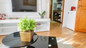 A modern living room with a flat-screen TV mounted on the wall, a small potted plant on a white console table, and a coffee machine in the kitchen area. - Apartamento aconchegante na Praça Pôr do Sol (São Paulo)