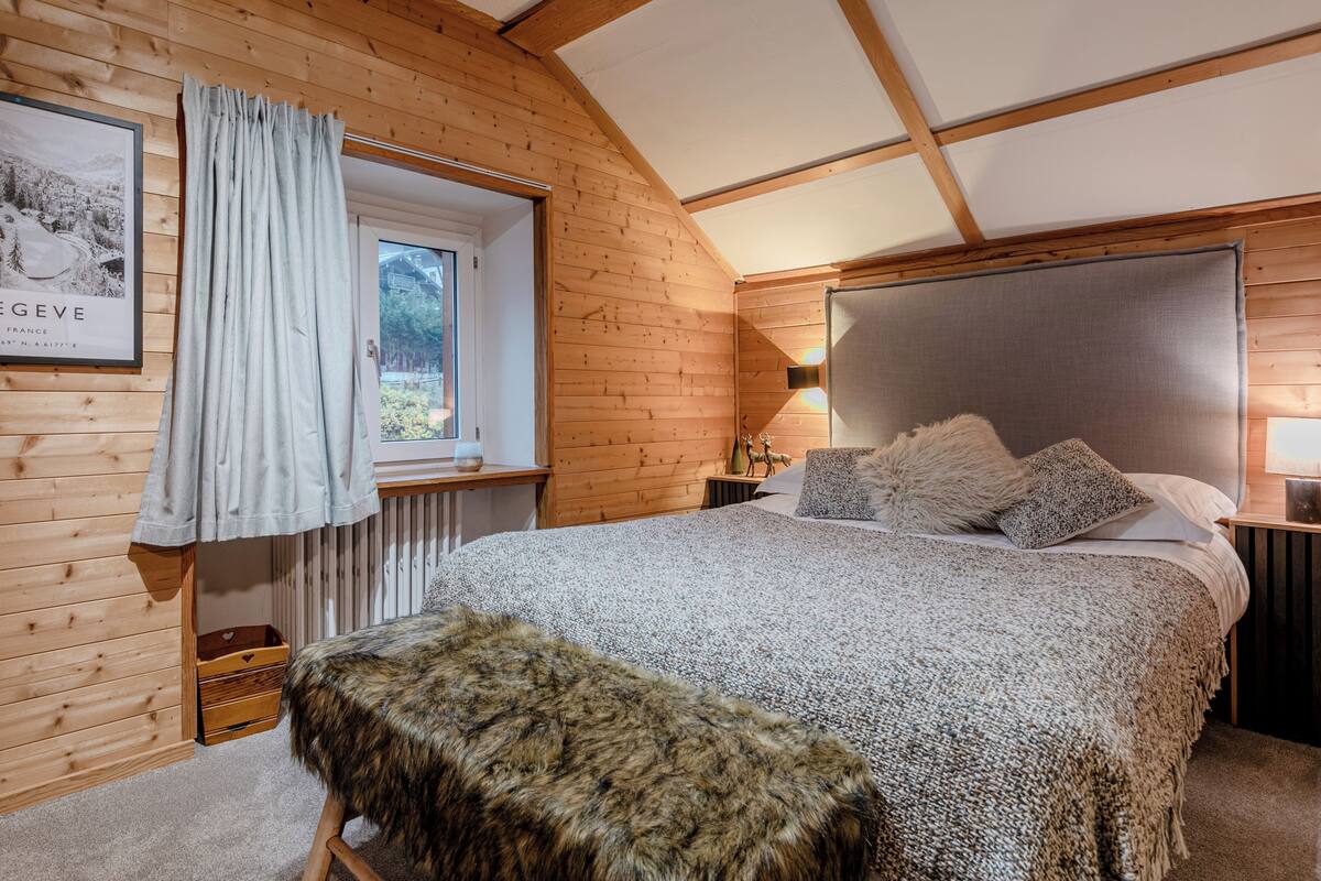 A cozy bedroom with wooden walls, a large bed with a fluffy blanket, a window with curtains, and a framed picture on the wall.