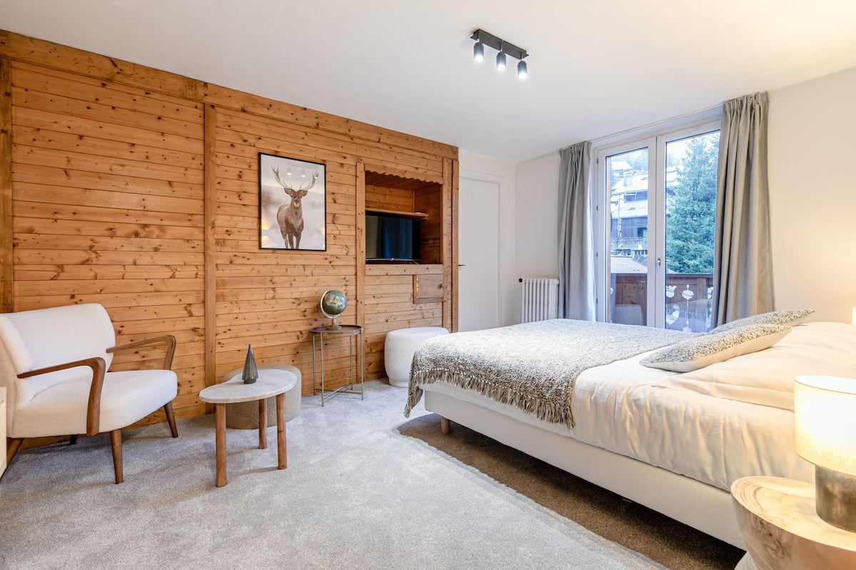 A bedroom with a wooden wall panel, a bed, a chair, a small table with a globe, and a window with curtains.