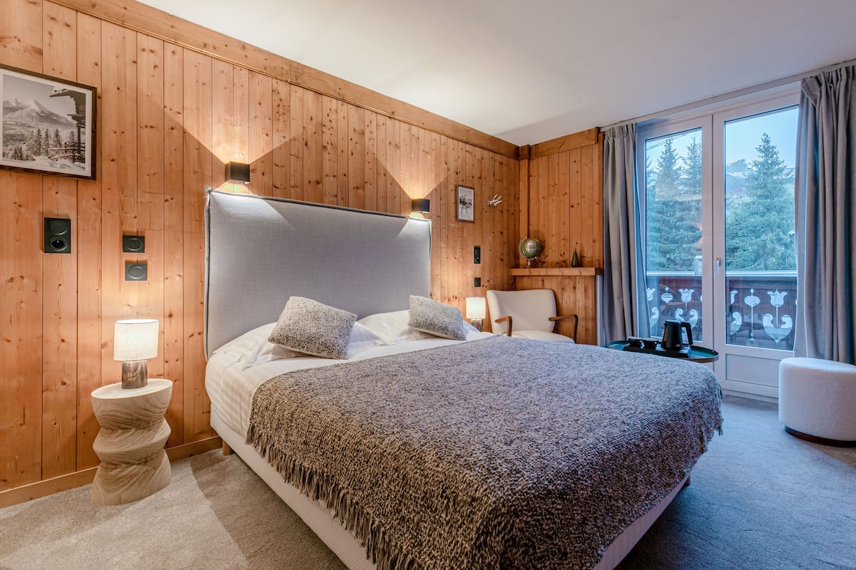 A wooden-paneled bedroom with a large bed, a nightstand with a lamp, a chair, and a view of trees through a window.