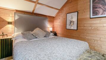A bedroom with a wooden ceiling, a bed with a grey and white patterned bedspread, a fluffy pillow, and a framed picture on the wall.