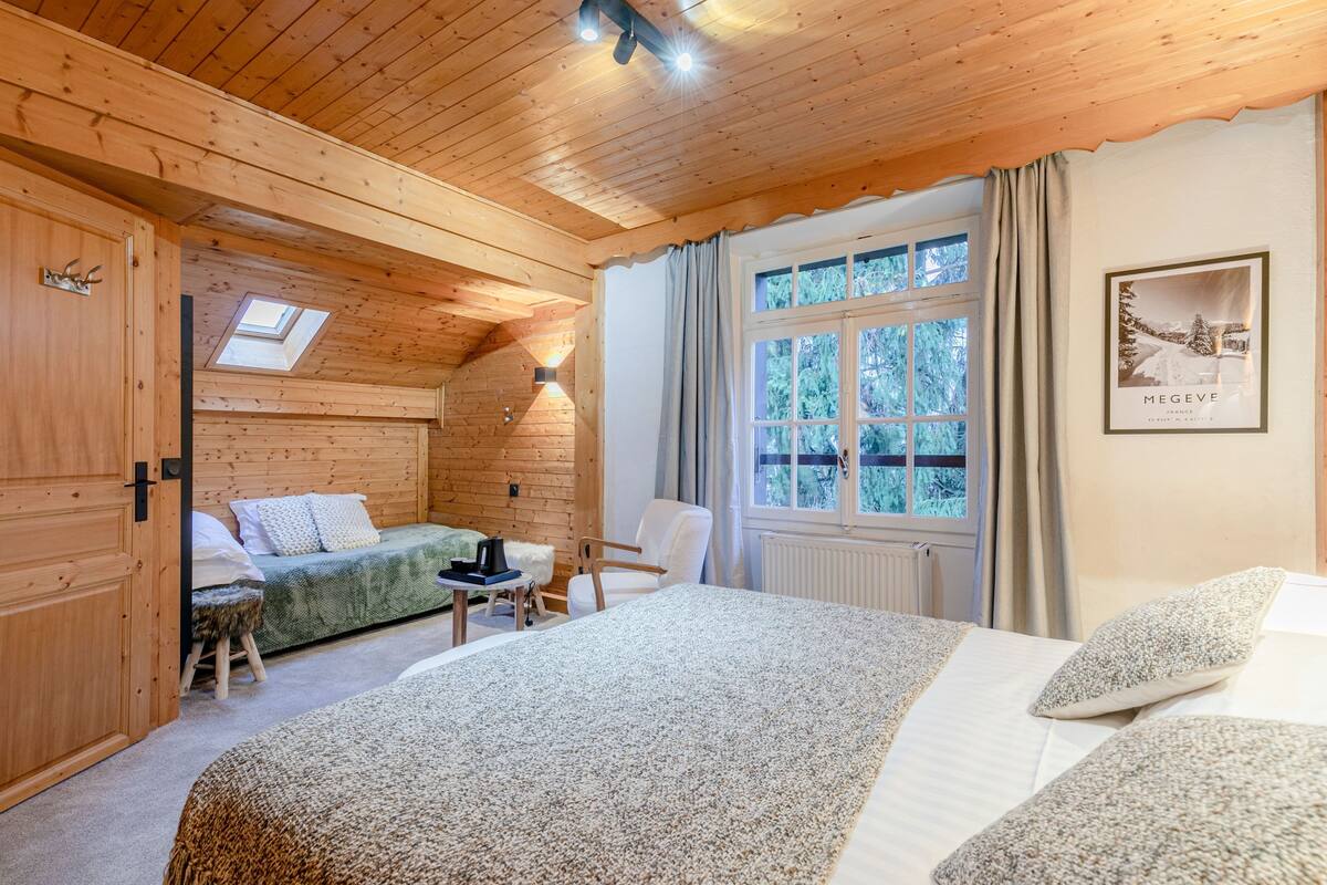A cozy log cabin room with a bed, a sofa, a chair, and a small table. There is a window with curtains and a framed picture on the wall.