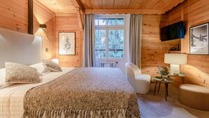 A cozy wooden cabin room with a bed, a chair, a small table with a lamp, and a television on the wall. - Le Chalet d'Antoine (Megeve)