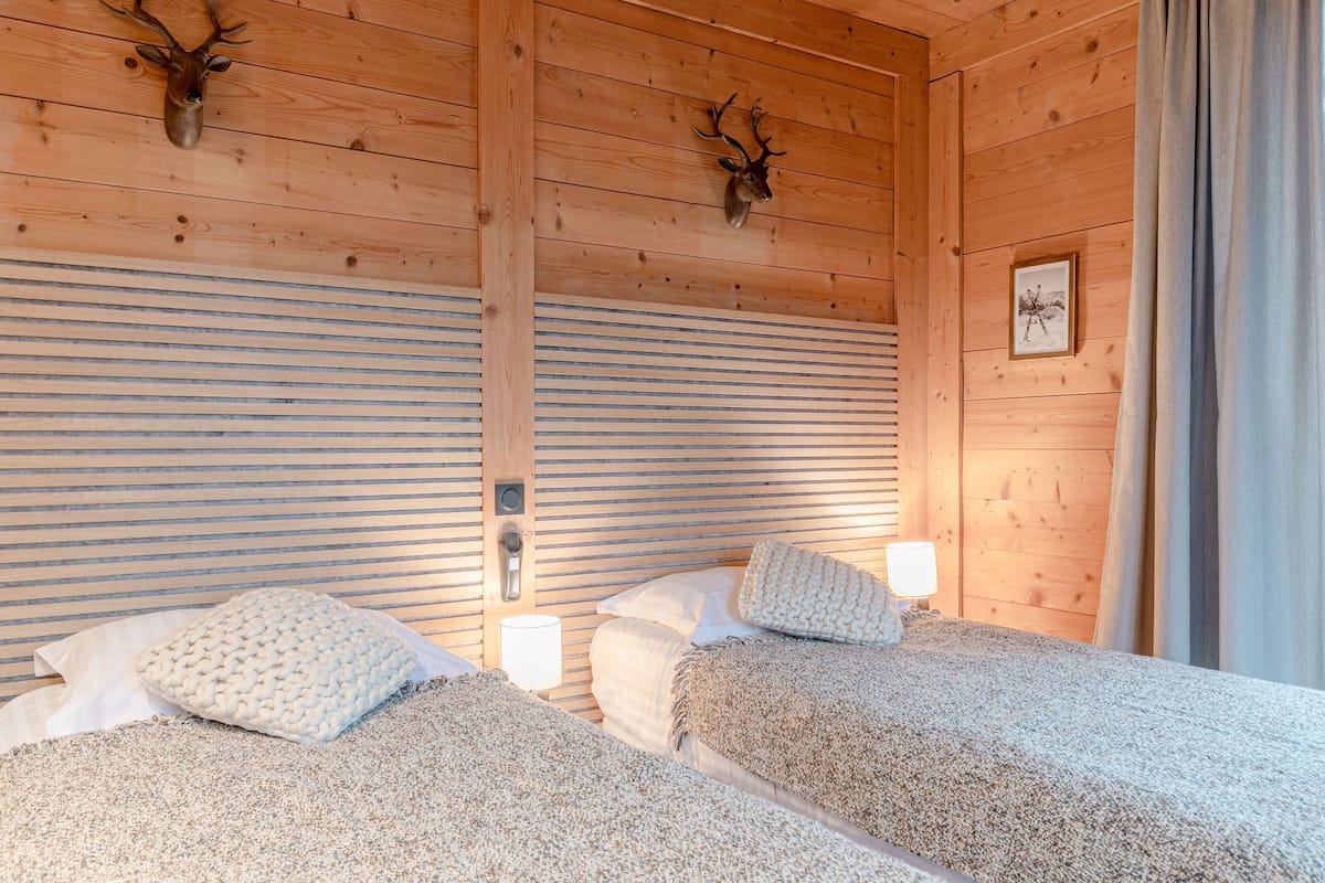 A wooden cabin room with two beds, a window with curtains, and deer antlers mounted on the wall.