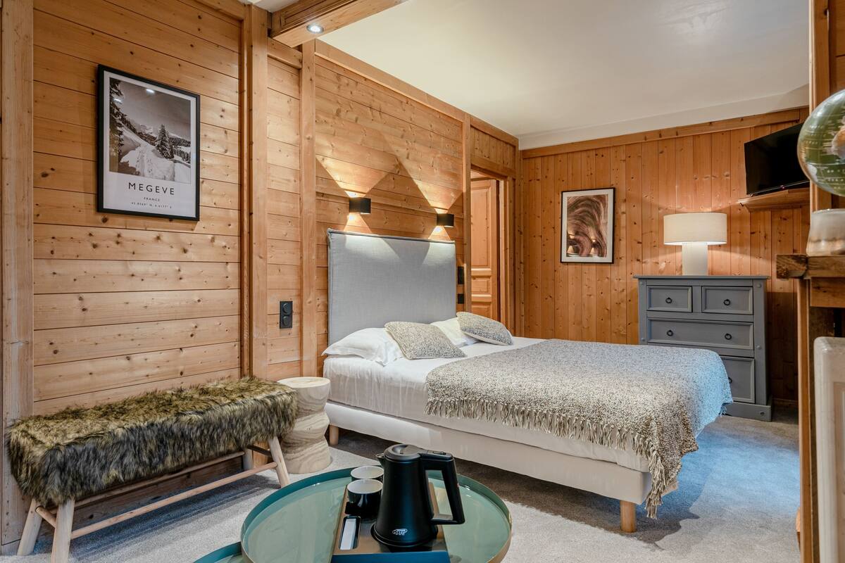 A wooden-paneled bedroom with a bed, a bench, a table with a kettle, and a framed picture on the wall.